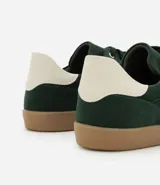 Championes de mujer estilo chuteira, color verde oscuro con detalles en blanco y suela de goma color caramelo. Presentan un diseño de corte bajo con cordones y un logo lateral en forma de arco.