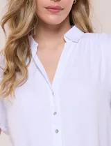 Camisa blanca de rayón con cuello camisero, escote en V y mangas cortas. Presenta abotonadura frontal con botones nacarados y detalles de tablas decorativas en el ruedo.