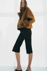 Trench corto color verde militar, con cuello solapa y capucha. Tiene manga larga con trabillas y botón en los puños, bolsillos delanteros y bajo asimétrico. Cierre frontal con botones. El tejido es repelente al agua.