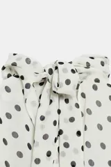 Blusa sin mangas blanca con estampado de lunares negros, cuello alto drapeado y nudo frontal en el bajo.