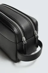 Neceser negro con textura grabada, cierre de doble cremallera y asa lateral. Interior con bolsillo con cremallera. Marca Mango.