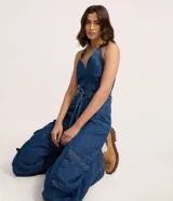 Mono largo de jean azul, sin mangas, con escote en V y cuello halter. Cuenta con cinturón de lazo en la cintura y bolsillos cargo en los laterales de las piernas.