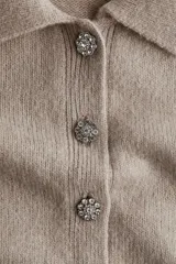 Saco de manga corta en punto suave color beige, con cuello camisero, cierre frontal de botones con strass y terminaciones acanaladas en mangas y bajo.