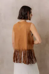 Chaleco de efecto gamuza en tono camel, sin mangas, con escote en V y diseño abierto. Presenta detalles de tachas metálicas, entramado artesanal en el frente y flecos largos en el ruedo.