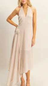 Vestido largo de tela fluida en color beige, con escote halter y detalle de flor aplicada en la cintura. Presenta un diseño asimétrico con caída suave y elegante.