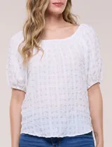 Blusa blanca de tela texturada y silueta fluida, con escote en U y frunces. Cuenta con mangas cortas abullonadas con elástico en los puños y ruedo redondeado.