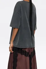 Camiseta de algodón orgánico color gris oscuro con efecto desgastado (acid wash). Presenta un corte oversize, escote redondo y un pequeño estampado gráfico en tono amarillo/verde en el pecho.