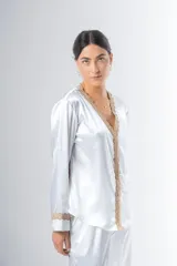 Pijama blanco de satén elastizado, compuesto por una camisa de manga larga con escote en V, abotonada adelante y con puntilla en escote y puños, y un pantalón recto con bolsillos laterales, elástico en la cintura y puntilla en el ruedo.