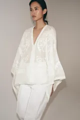 Blusa tipo capa de algodón con escote en pico y mangas largas amplias. Presenta bordados florales a tono en toda la prenda, bajo asimétrico y cierre frontal con botones.