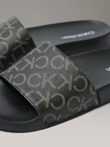 Ojotas Calvin Klein con diseño de monograma de la marca en color beige sobre fondo negro. Presentan una suela anatómica contorneada para mayor confort y una tira superior de material sintético.