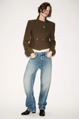 Chaqueta corta de paño color verde militar, con cuello subido, manga larga con puños abotonados y cierre frontal cruzado con botones metálicos. Presenta hombreras y bolsillos de vivo en el delantero.