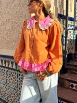 Camisa de manga larga color naranja con cuello grande en contraste color rosa y detalle de volados en el borde. Presenta un diseño con botones frontales y un detalle de tela rosa en la parte inferior.