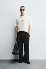 Camiseta boxy fit de color blanco con cuello redondo y mangas cortas. Presenta un parche rectangular en el centro del pecho con texto impreso y costuras decorativas a contraste en los laterales.