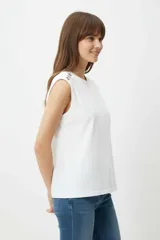 Remera de algodón blanca, de corte holgado y sin mangas, con cuello a la base y detalle de ojalillos metálicos sobre los hombros.