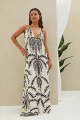 Vestido largo color blanco con estampado de palmeras en tonos negros y dorados, con escote halter y diseño fluido sin mangas.