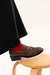 Mocasín de cuero color chocolate con acabado texturizado y diseño enrejillado en la capellada. Presenta horma cuadrada, taco bajo y suela de goma.