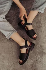 Sandalia de cuero color tostado con tiras anchas, tira en el talón y hebilla metálica rectangular ajustable. Cuenta con suela de goma con tacón bajo y plataforma.