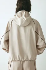 Campera rompevientos de color beige, modelo oversized con cuello alto y capucha con cordón ajustable. Cuenta con cierre frontal marrón, mangas raglán con puños elásticos, bolsillos laterales y detalles de ribetes en tono marrón oscuro.