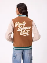Campera estilo varsity con cuerpo en color marrón y mangas en tono beige. Presenta cierre frontal con botones a presión, cuello, puños y ruedo con franjas tejidas en verde y blanco. Incluye un parche con la letra R en el pecho y detalles bordados en las mangas.