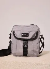 Bandolera Rusty modelo Vansi, color gris claro, con bolsillo frontal con solapa y cierre de hebilla plástica. Incluye correa ajustable y desmontable en color negro y el logo de la marca impreso en el frente.