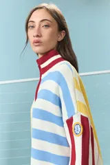 Sweater de punto de algodón con diseño de bloques de rayas verticales y horizontales en colores rojo, amarillo, azul y blanco. Presenta cuello alto con cierre frontal parcial y mangas largas con puños acanalados.