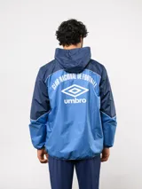 Campera de lluvia Umbro del Club Nacional de Football, con diseño de bloques de color en rojo y azul marino. Presenta capucha, cierre frontal, puños elásticos y detalles de piping blanco. Incluye logo de Umbro y escudo del club en el pecho.