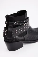 Botín de cuero vacuno negro con textura grabada, taco cuadrado y punta fina con detalle metálico. Presenta una tira decorativa con tachas metálicas y hebilla ajustable alrededor del tobillo.