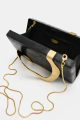 Bolso tipo clutch negro de formato rectangular con diseño rígido, adornado con un detalle metálico dorado de líneas onduladas en el frente. Cuenta con un cierre imantado y una correa de cadena metálica fina y dorada para llevar al hombro o cruzada.