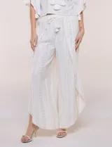 Pantalón palazzo cruzado color blanco con trama en hilo metalizado lurex, cintura elastizada con cordón ajustable con borlas y diseño de piernas anchas con bordes de puntilla y tajo profundo en el frente.