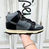 Championes Nike Dunk High de corte alto, con diseño en combinación de cuero negro y gris. Presenta suela de goma en tono crema envejecido y detalle de dije metálico colgante en el cuello.