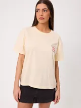 Remera blanca de corte holgado, con cuello redondo y mangas cortas. Presenta un pequeño estampado en el pecho con diseño floral y tipografía.