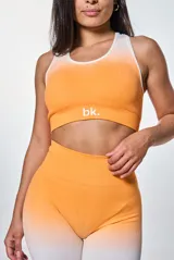 Conjunto deportivo de dos piezas compuesto por un top de cuello redondo y un short de tiro alto, ambos con diseño degradado en tonos naranja y blanco. El top presenta el logo 'bk.' estampado en el frente.