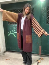 Sacon de diseño asimétrico en tonos bordó y marrón, con una manga lisa y otra a rayas horizontales. Presenta bolsillos frontales con apliques de cuero sintético y corte largo abierto.