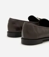 Mocasín femenino de cuero sintético color marrón oscuro, con punta redondeada y suela plana. Presenta un diseño con tiras cruzadas en el empeine decoradas con ojales metálicos y una hebilla ajustable.