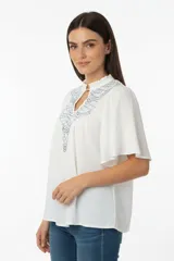 Blusa de viscosa color blanco, con cuello en V y detalle de bordado geométrico en tonos azules en la parte frontal. Presenta mangas cortas con vuelo y un corte holgado.