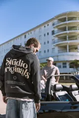 Canguro negro con capucha, de corte holgado y mangas raglán. Presenta un gran estampado en color beige en la espalda con la inscripción "Studio Originals" y texto descriptivo, además de puños y dobladillo acanalados.