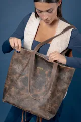 Bolso tipo tote grande de cuero con acabado texturizado y efecto desgastado en tonos marrones. Presenta detalles de costuras trenzadas en los laterales, doble asa de hombro y una placa metálica con el logo de la marca en el frente.
