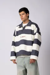Buzo tipo polo de corte boxy, con diseño de franjas horizontales anchas en color verde oscuro y crema. Presenta cuello camisero, cierre frontal parcial y detalles de costuras desflecadas. Incluye logo bordado en el pecho.