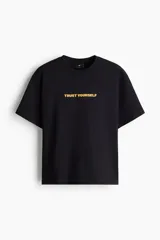 Remera negra de algodón con cuello redondo, mangas cortas y hombros caídos. Presenta un estampado frontal con la frase "TRUST YOURSELF" en color amarillo.