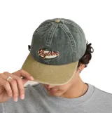 Gorra tipo baseball con corona en color verde lavado y visera en color beige. Presenta un parche bordado frontal con la palabra 'Rhythm' en tipografía cursiva.