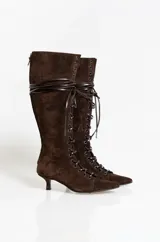 Bota alta de gamuza color marrón oscuro, con diseño de cordones frontales y cierre lateral. Presenta una silueta estilizada con punta fina y un taco bajo tipo kitten heel.