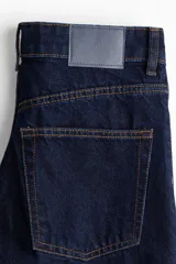 Jean de denim azul oscuro rígido, con diseño de pierna amplia y tiro alto. Presenta bolsillos, pinzas delanteras y traseras, y un dobladillo ancho vuelto hacia arriba en los tobillos.
