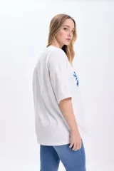 Remera blanca de corte amplio con estampado de palmeras, cabaña y aves en color azul.