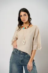 Blusa de lino color beige con cuello bote y mangas cortas con detalle de lazos laterales y bordes desflecados. Presenta botones de nácar en el frente.