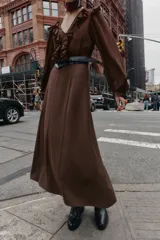 Vestido midi de color marrón con diseño combinado, escote en pico con volantes, mangas largas con puños elásticos y falda evasé. Incluye un cinturón negro con hebilla dorada en la cintura y cierre lateral con cremallera oculta.