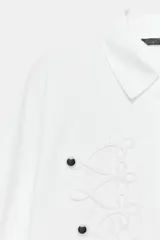 Camisa de popelín blanca con cuello solapa y manga larga. Presenta un diseño frontal con bordados en relieve y botones decorativos negros. Cierre frontal con botones ocultos.