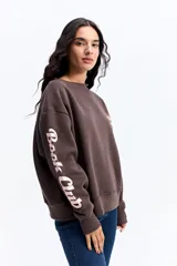 Buzo de algodón color marrón chocolate, con cuello redondo y diseño holgado. Presenta una estampa gráfica en la espalda con motivos de atardecer y montañas, junto con texto decorativo. Además, incluye una estampa en la manga con letras grandes.