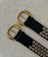 Cinturón negro de cuero ecológico con hebilla metálica dorada y doble hilera de ojales dorados a lo largo de toda la tira.