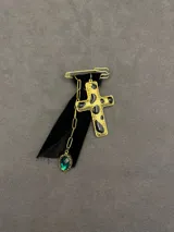 Pin decorativo con forma de cruz dorada de estilo vintage, adornada con piedras incrustadas. La pieza incluye una base de tela de terciopelo desflecada, una cadena metálica y un pequeño dije colgante.