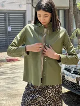 Camisa de cuero color verde oliva, con cuello camisero, cierre frontal mediante broches metálicos y mangas largas con puños abotonados.
